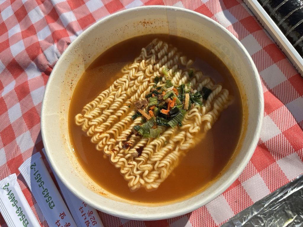 Close-up of Korean instant ramen in spicy broth with vegetable toppings, served in a paper bowl on a red checkered picnic mat at Banpo Hangang Park in Seoul.