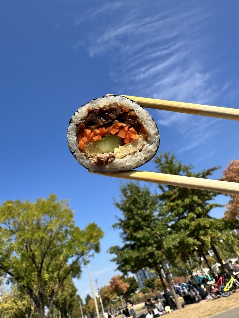 Close-up of a Korean gimbap slice held with chopsticks against a bright blue autumn sky at Banpo Hangang Park in Seoul.