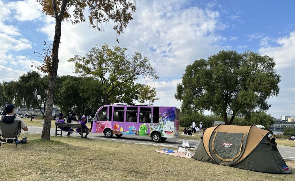 Colorful purple park shuttle vehicle with cartoon characters passing picnic tents and visitors at Banpo Hangang Park in Seoul.